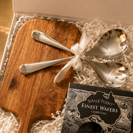 Wooden cutting board with silver utensils and a box of Shale Point Finest Wafers Charcoal on a textured surface.