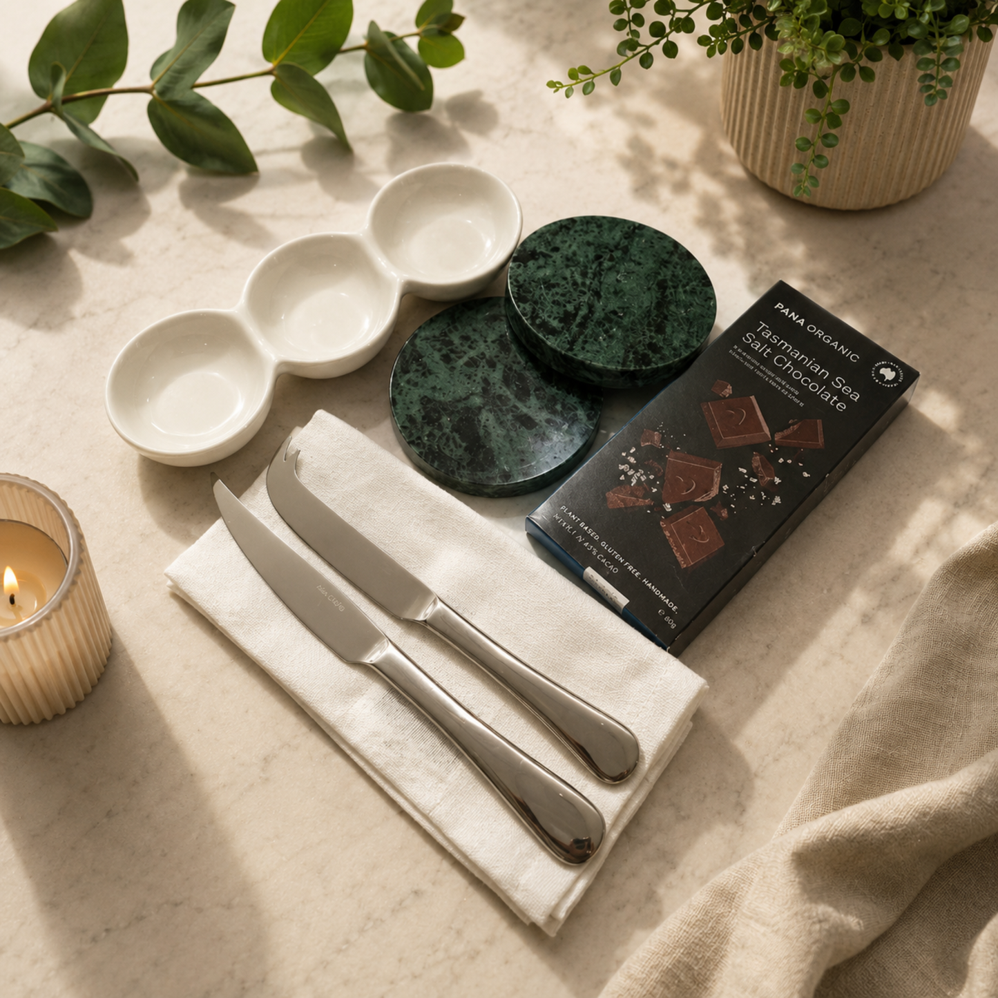 Table setting with ceramic bowls, knives, a box of chocolates, and a candle on a light surface.