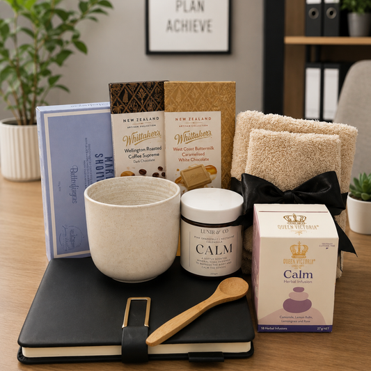 Assorted items including a mug, candles, and a notebook on a desk with a plant and framed picture in the background.