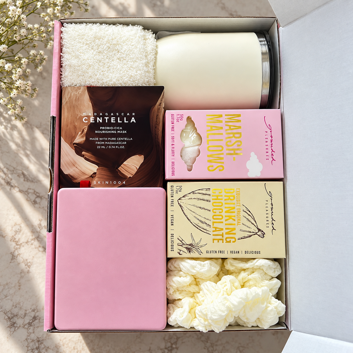 An open pink gift box arranged with skincare and beauty items on a marble surface: a white textured towel, a white candle, a brown sheet mask package labeled Centella, a pink marshmallow mask package, a beige chocolate mask package, a pink tin, and two white scrunchies.