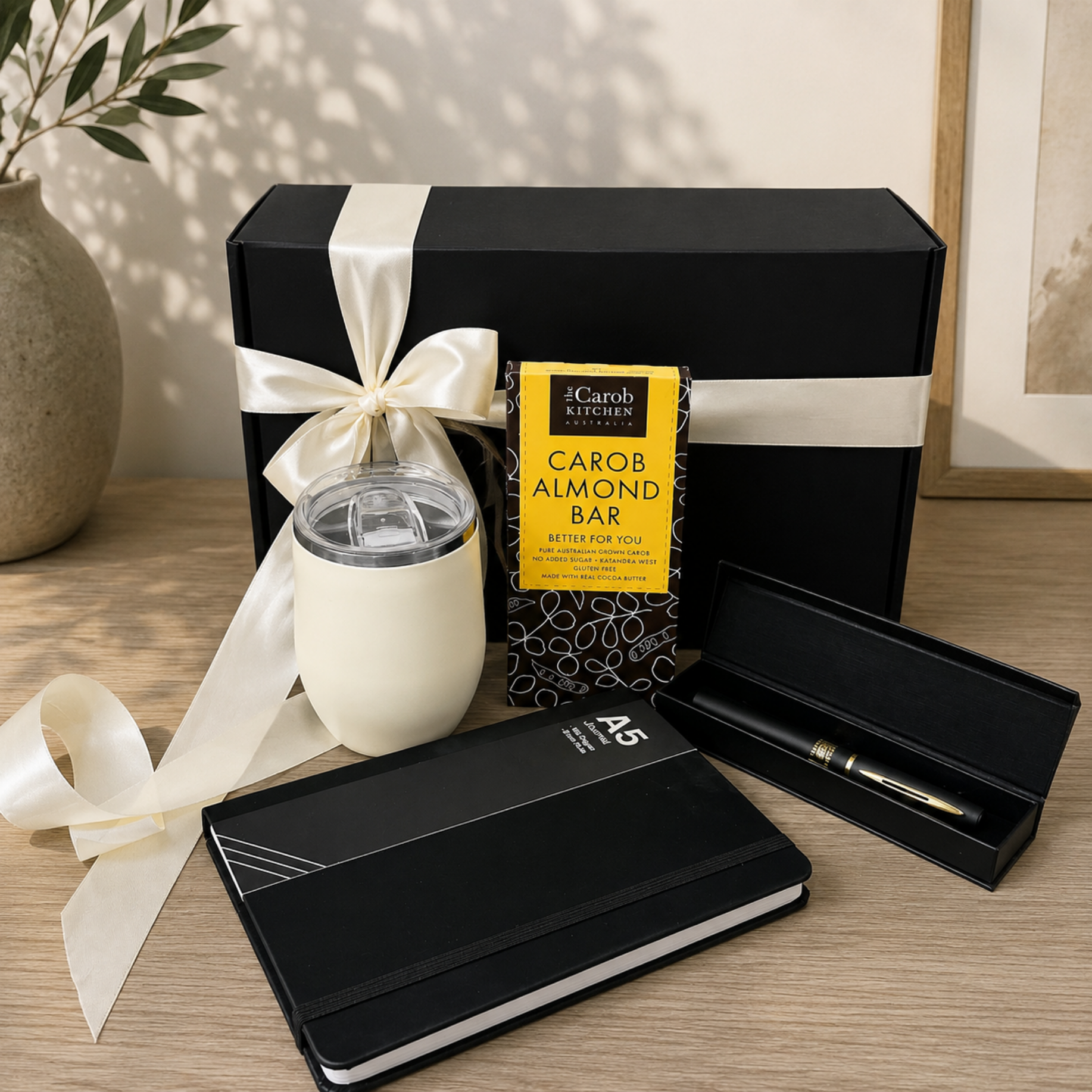 A curated gift set on a wooden table featuring a black gift box with a cream satin ribbon, a black notebook, a black pen in an open case, a cream insulated tumbler, and a yellow box of Garob Kitchen Carob Almond Bar.