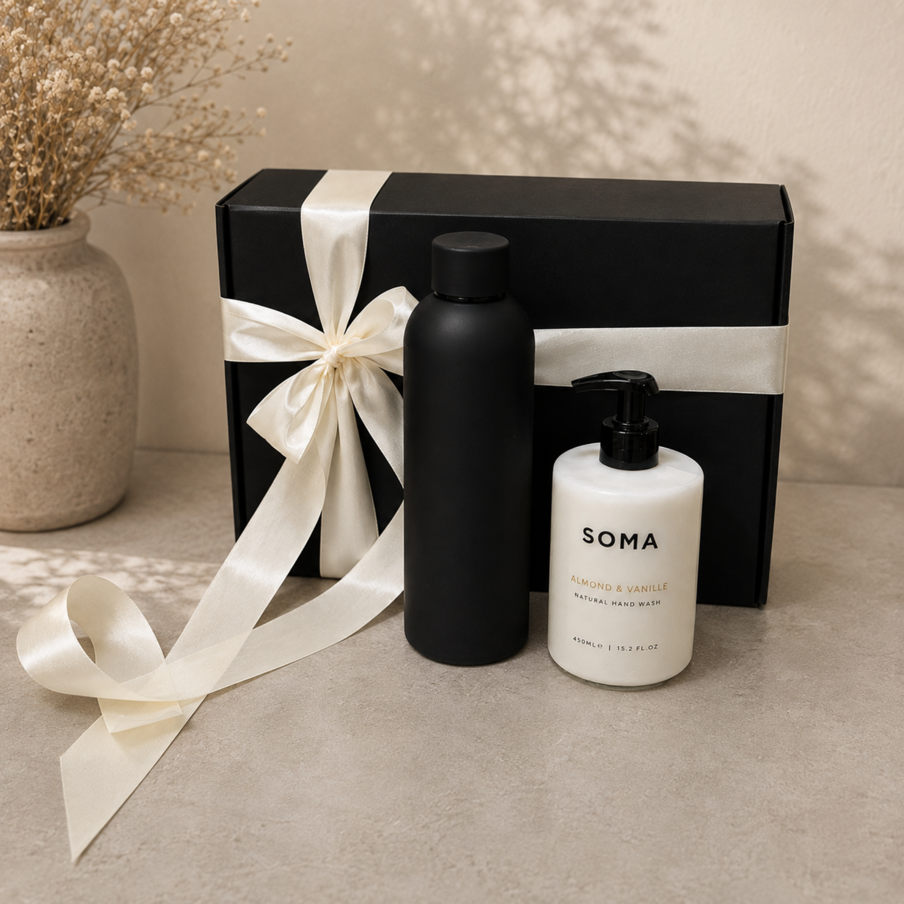 Soma natural hair wash gift set featuring a matte black bottle and a white pump bottle with black cap, presented in front of a black gift box tied with a cream satin ribbon, on a beige stone surface.