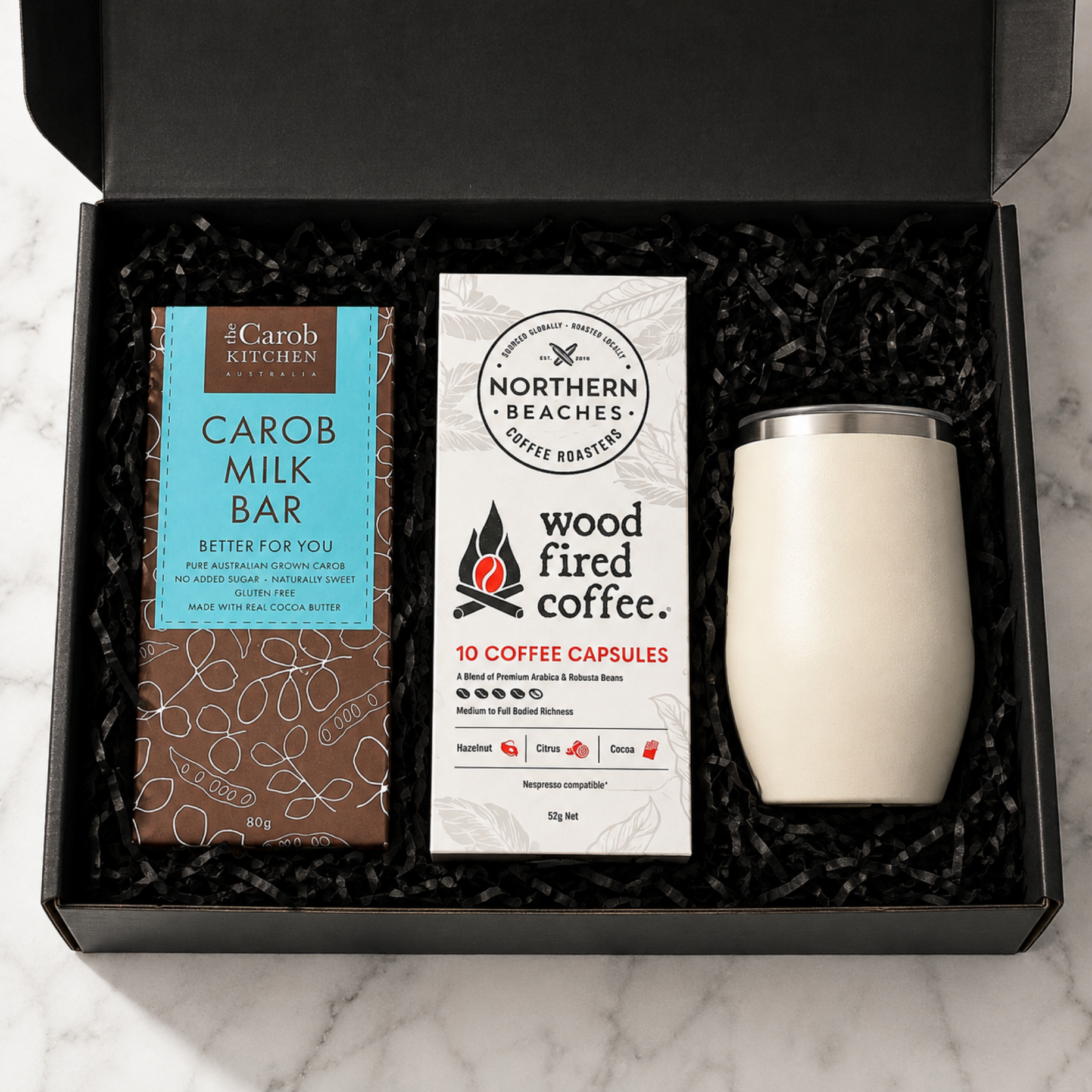 A curated coffee gift set in an open black box with black shredded filler, featuring a bar of carob milk bar, a box of Northern Beaches wood fired coffee capsules, and a cream-colored insulated tumbler with a silver lid.