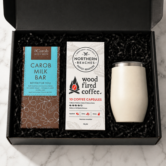 A curated coffee gift set in an open black box with black shredded filler, featuring a bar of carob milk bar, a box of Northern Beaches wood fired coffee capsules, and a cream-colored insulated tumbler with a silver lid.