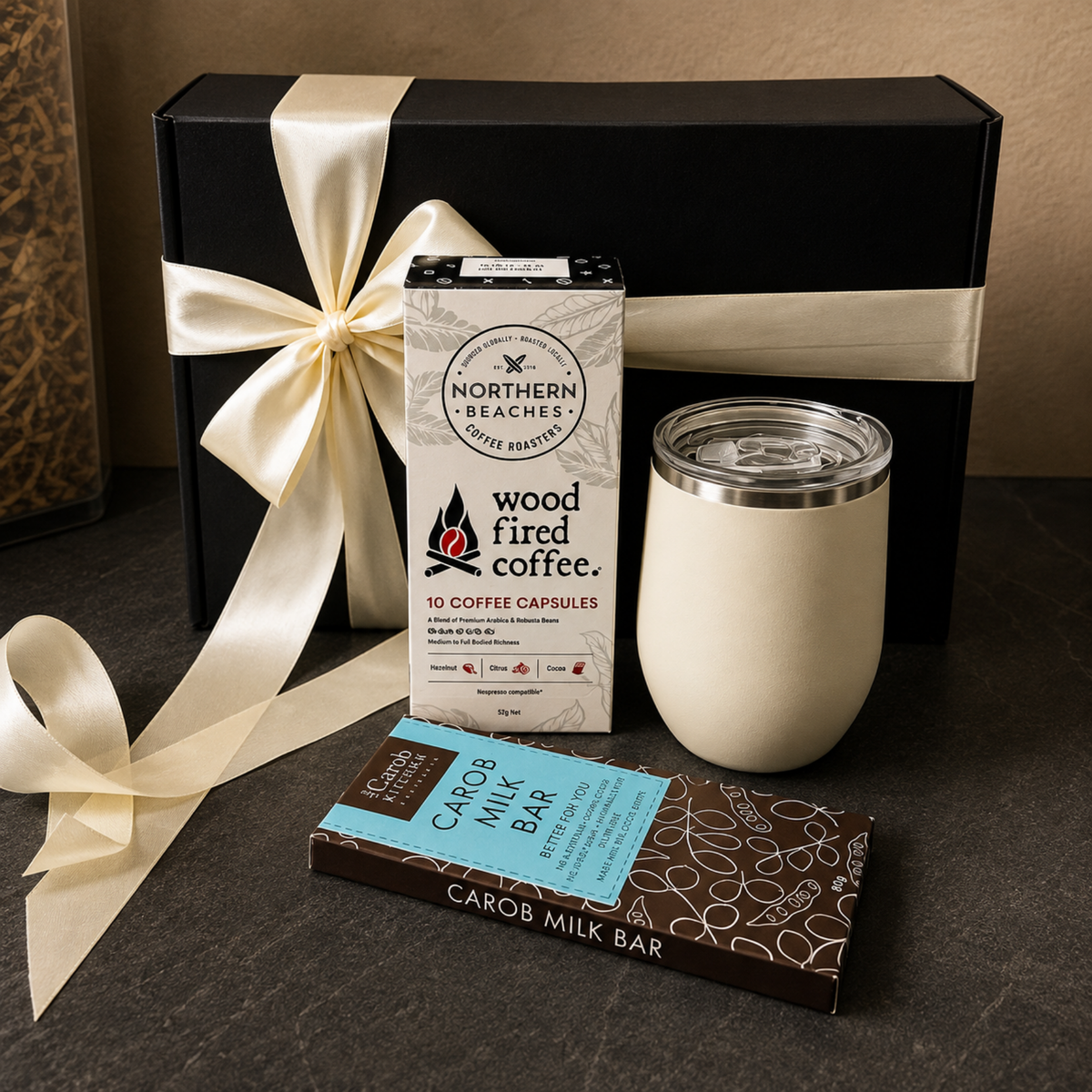 A curated gift set on a dark stone surface featuring a black gift box with a cream ribbon, a Northern Beaches coffee capsule box, a cream insulated tumbler, and a Carob Milk Bar chocolate bar.