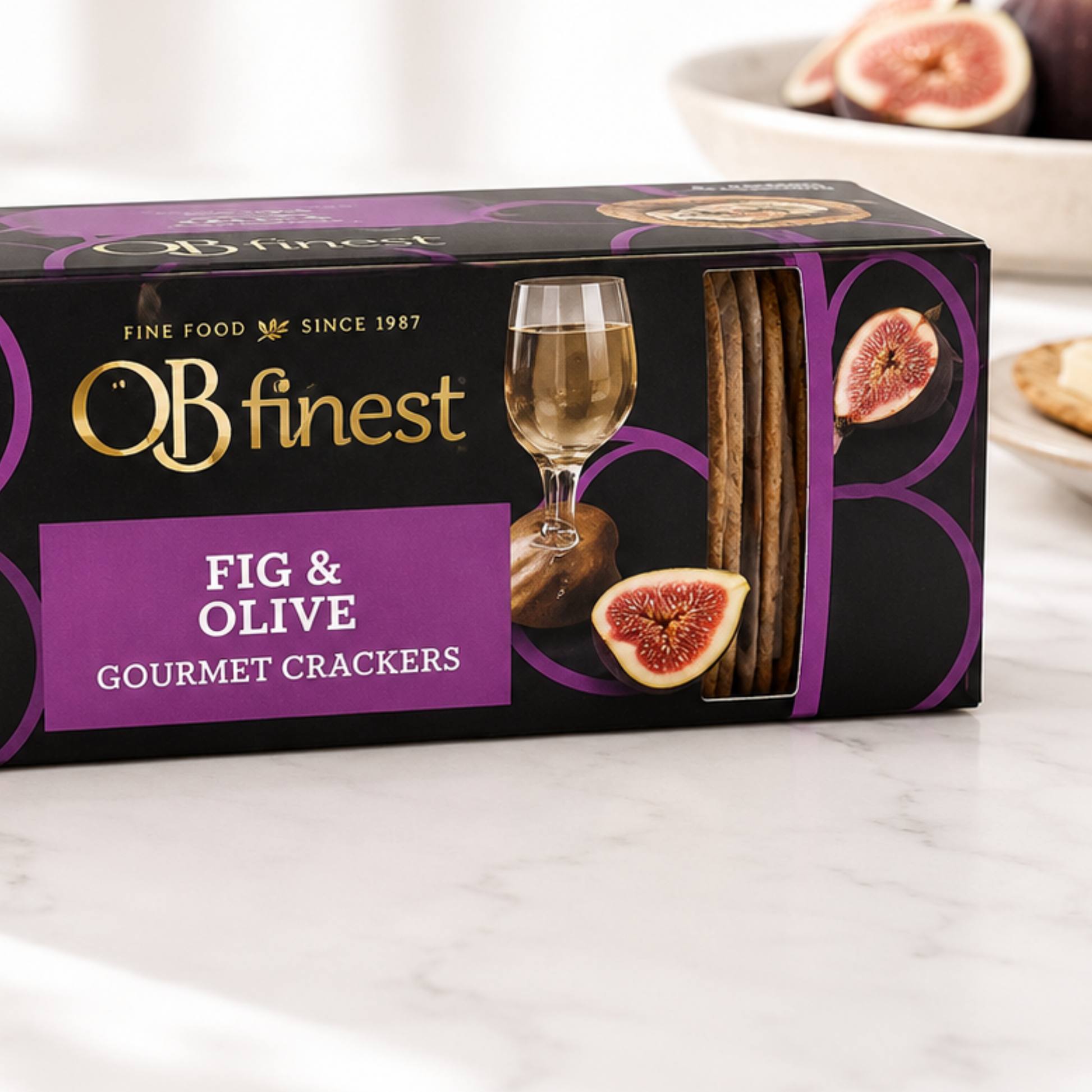 Box of OB finest Fig & Olive Gourmet Crackers on a marble surface with figs and a glass of wine in the background.