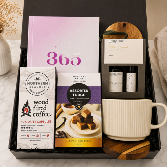 A curated coffee and tea gift set arranged in a black presentation box on a marble surface. Visible items include a pink and purple book, Northern Beaches 'wood fired coffee' capsules, Millicent Grove assorted fudge boxes, Herbalist Tea 'Falsely Indulgent' tea, two Herbalist Pause supplement bottles, and a white ceramic mug on wooden trivets.