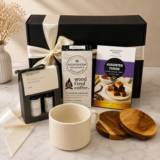 A coffee gift set arranged on a marble surface with a large black gift box tied in cream ribbon behind it. In front are a cream ribbed mug, a package of Northern Beaches wood fired coffee capsules, a package of Millcent Curl assorted fudge, a Herbalist Grove Passion Citrus tea box, two small spice jars, and wooden coasters.