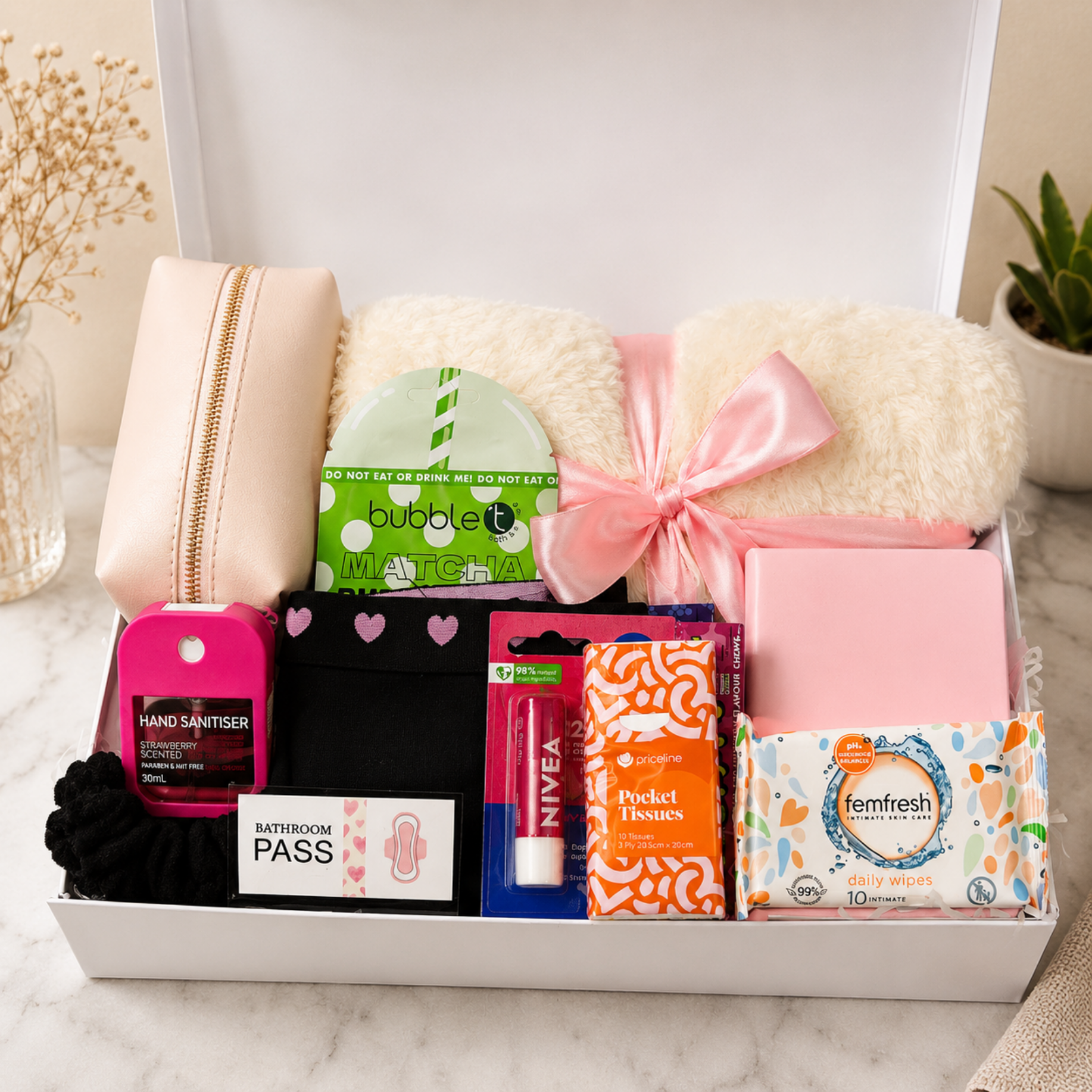 A curated gift box set containing a beige zippered pouch, two cream faux-fur pillows tied with a pink ribbon, a pink hand sanitizer bottle, black socks, a pink lip balm tube, orange patterned toilet paper, a box of Temfresh wipes, and a bathroom pass, all arranged in an open white box on a marble surface.