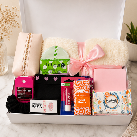 A curated gift box set containing a beige zippered pouch, two cream faux-fur pillows tied with a pink ribbon, a pink hand sanitizer bottle, black socks, a pink lip balm tube, orange patterned toilet paper, a box of Temfresh wipes, and a bathroom pass, all arranged in an open white box on a marble surface.