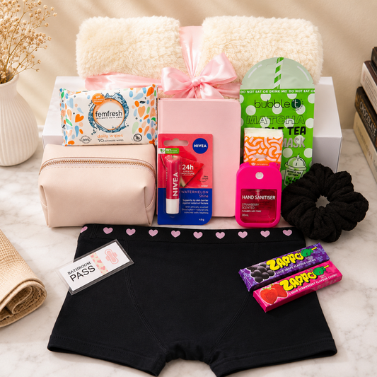 A curated gift set arranged on a light marble surface, featuring black heart-trimmed underwear, a pink gift box with a pink bow, folded cream towels, a blush makeup pouch, a Tenfresh soap pack, a Nivea lip balm tube, a bubble wrap bag, a pink hand sanitizer bottle, a black scrunchie, two packs of Zippy candy, and a folded beige towel. No people or real-life environment are present, classifying this as Product/Packaging.
