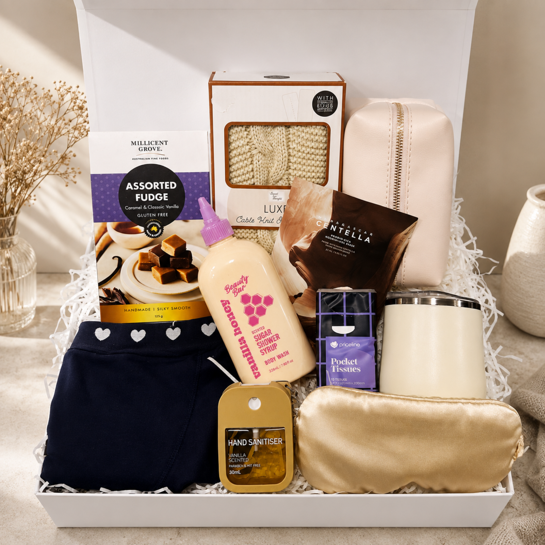 A curated gift box arrangement featuring a navy blue pair of underwear with white hearts, a beige satin zip pouch, a beige satin sleep mask, a hand sanitizer bottle, a boxed towel, a boxed candle, a boxed chocolate bar, and a box of Millicent Grove Assorted Fudge, all arranged in white packing paper inside an open white gift box.