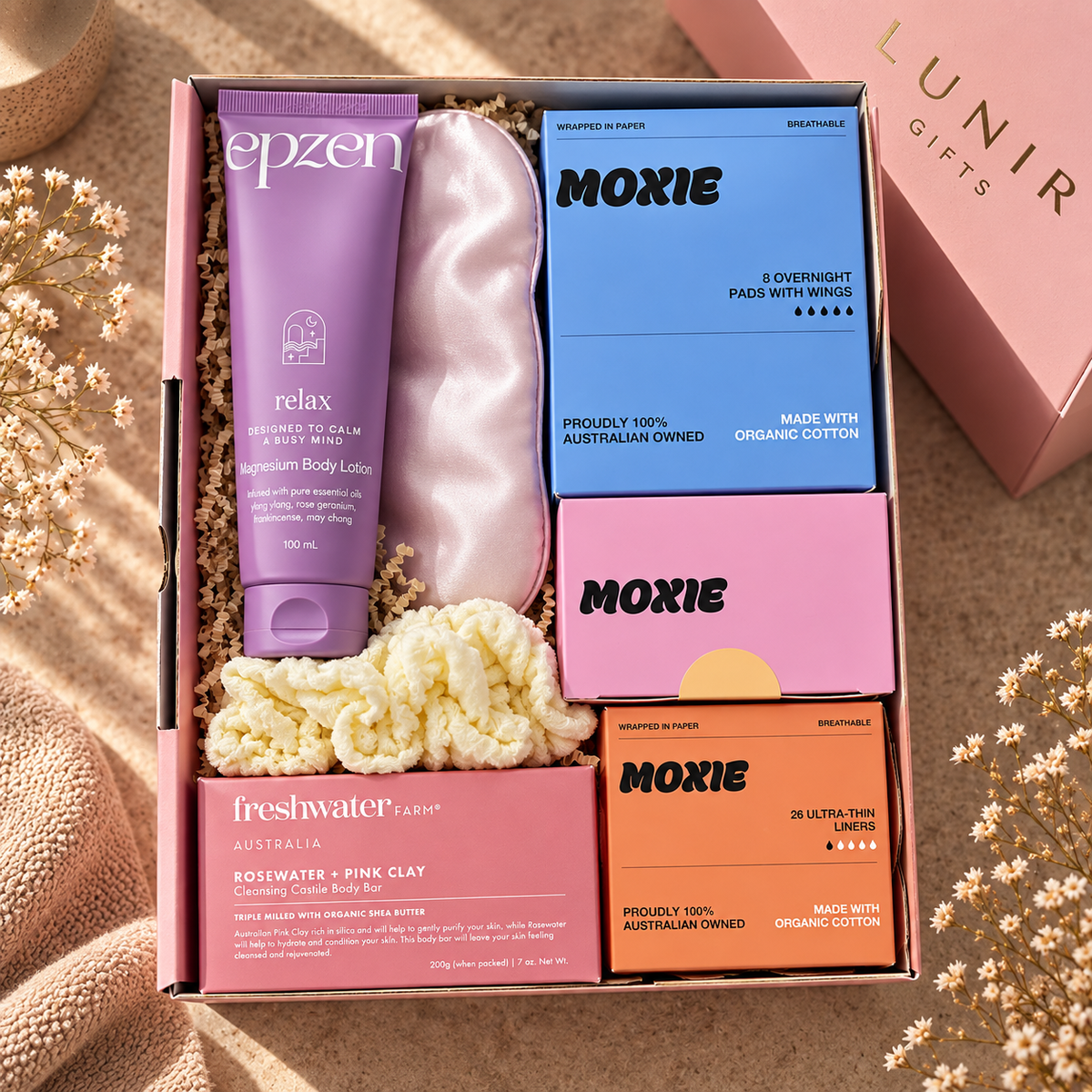 A curated wellness gift box set from Epzen, Moxie, and Freshwater Farm, arranged in a pink box with shredded paper filler. The set includes a purple Epzen Relax body lotion tube, a pink satin eye mask, a blue Moxie 8 Overnight Pass with Mint, a pink Moxie box, a pink Freshwater Farm Rosewater + Pink Clay body wash box, and an orange Moxie box, all styled on a beige fabric background with dried flowers.