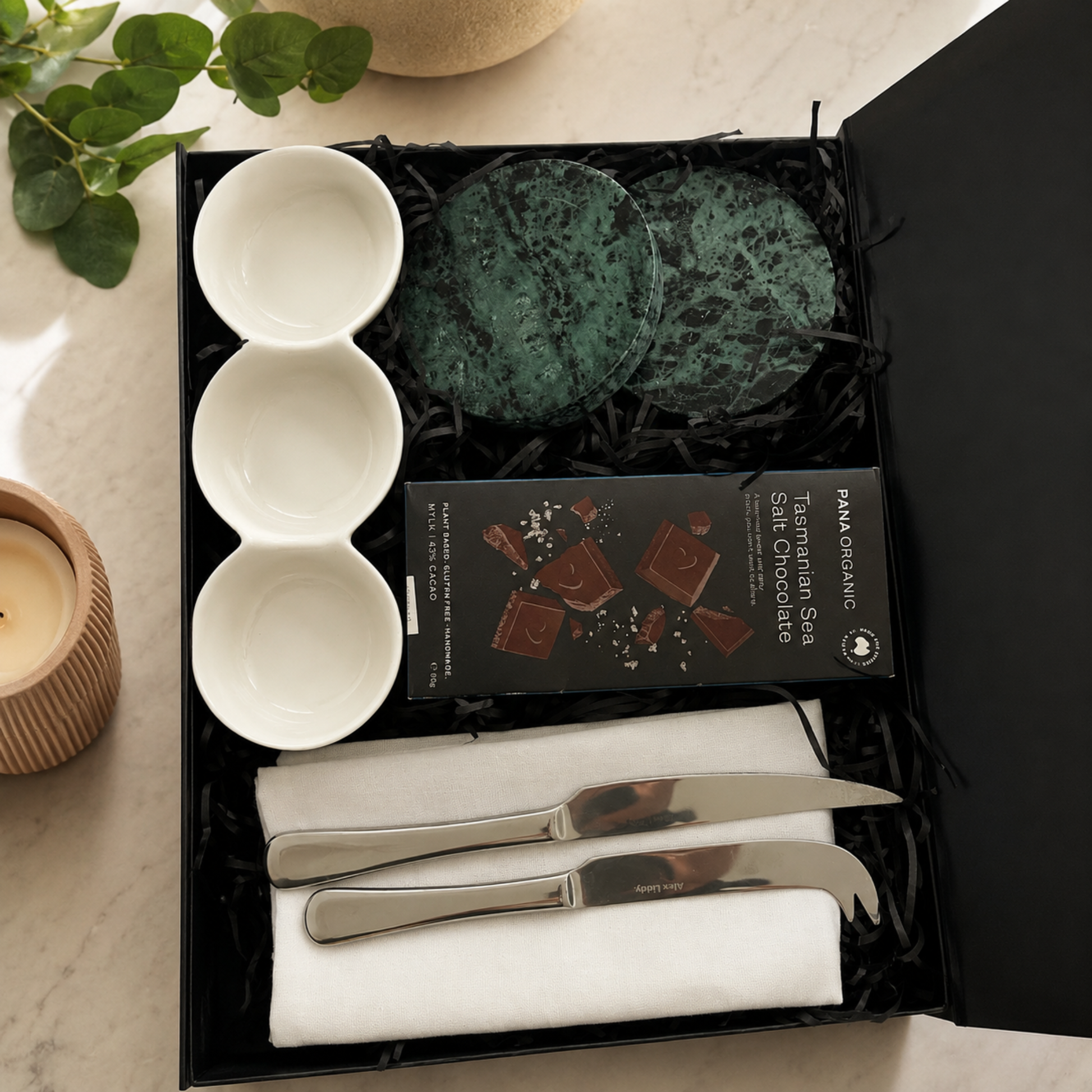 Gift set with coasters, chocolate, and cutlery in a black box on a light surface.