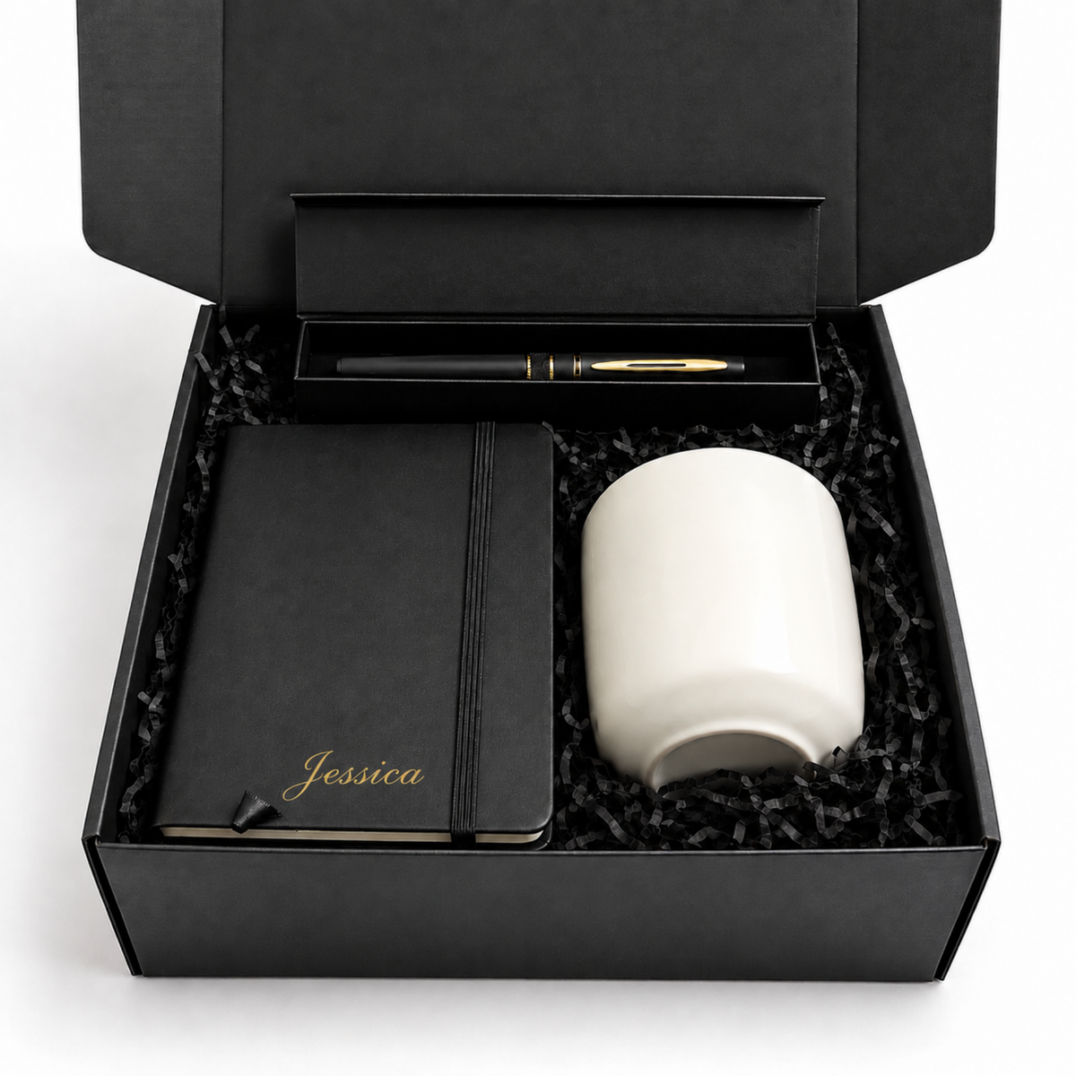 A black gift box with shredded black paper filler contains a black notebook with gold 'Jessica' script, a white ceramic mug, and a black pen with gold trim in a matching black tray. The box lid is open, photographed on a white background.