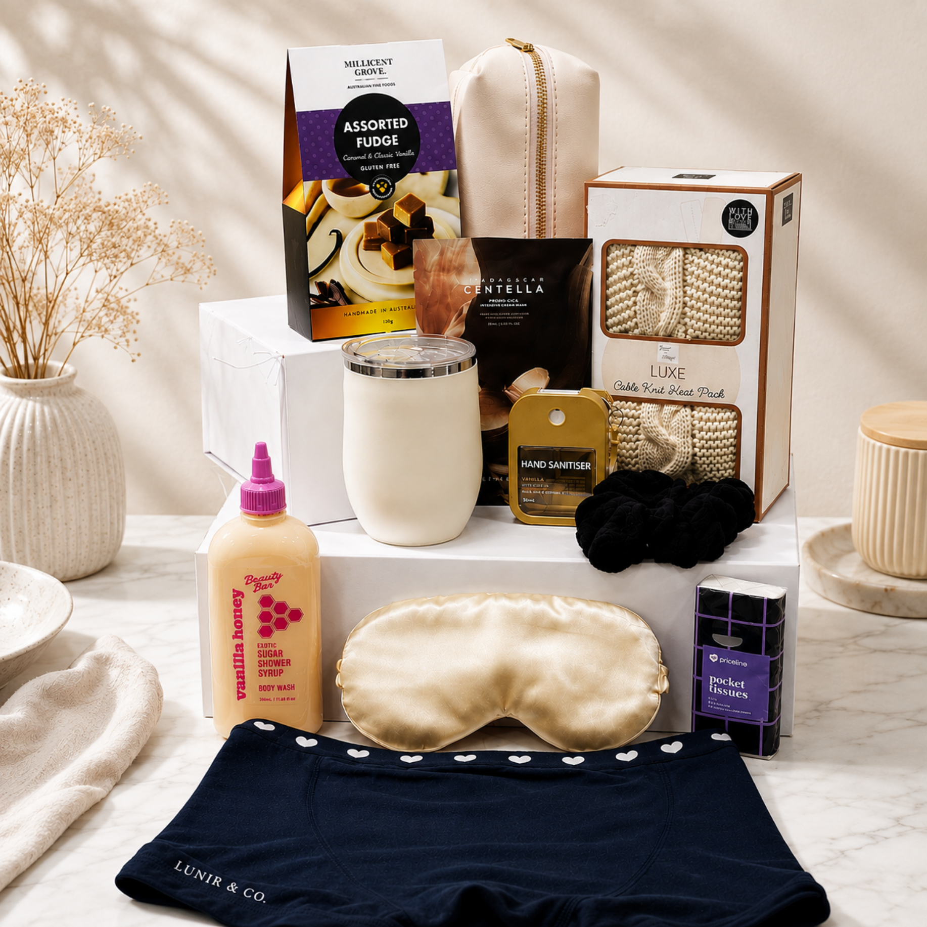 A curated gift set arranged on white display blocks and a marble surface, featuring a satin sleep mask, navy underwear, a tumbler, boxed socks, a bottle of vanilla honey soap, a candle, a pouch, and other wellness/luxury items. Visible brands include Lunir & Co on the underwear and tumbler, Luxe on the sock box, and Vanililla Honey on the soap bottle.