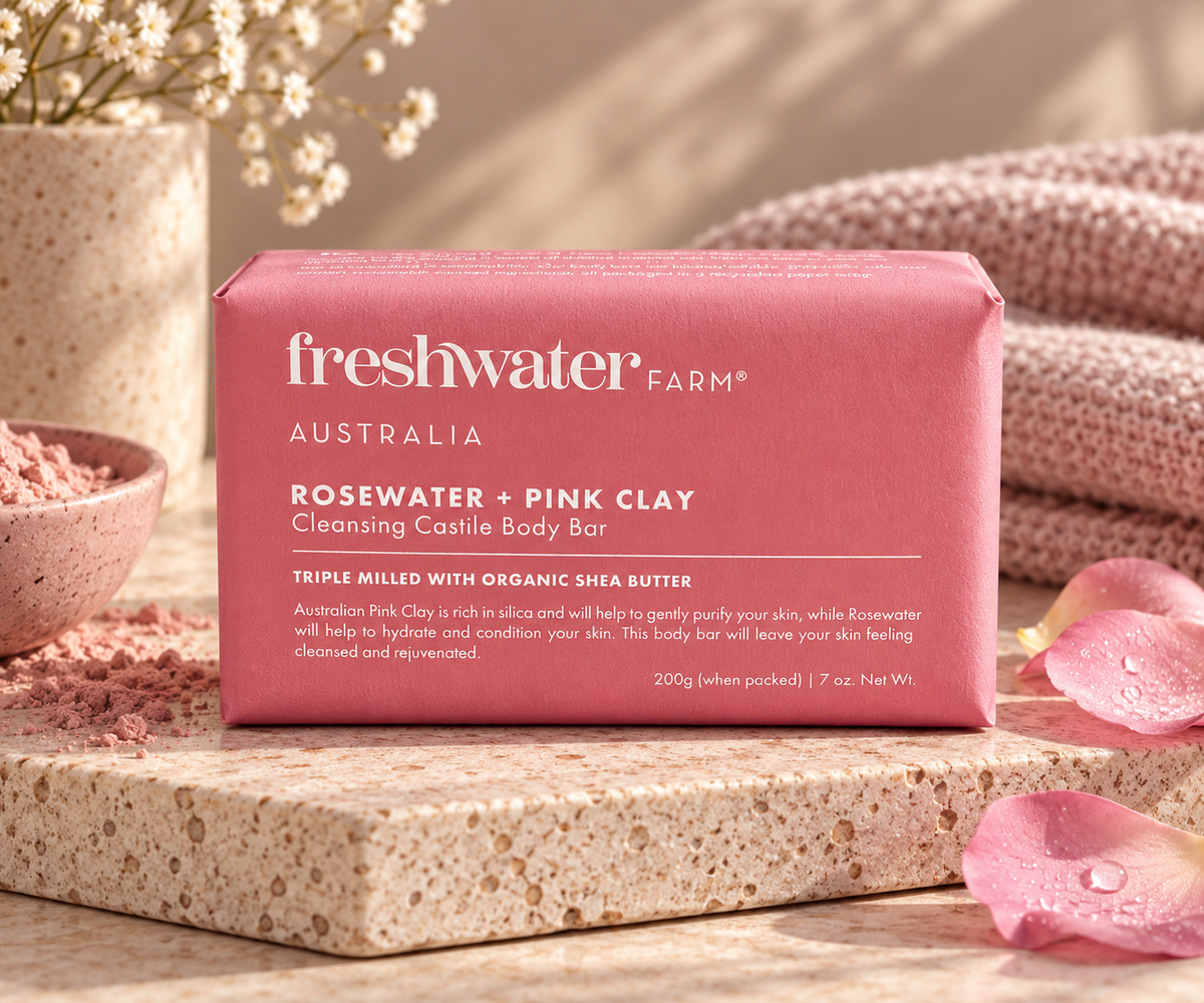 A pink rectangular bar of Freshwater Farm Rosewater + Pink Clay Cleansing Castile Body Bar, displayed on a stone slab with pink clay powder, a pink towel, and dried flowers in the background.