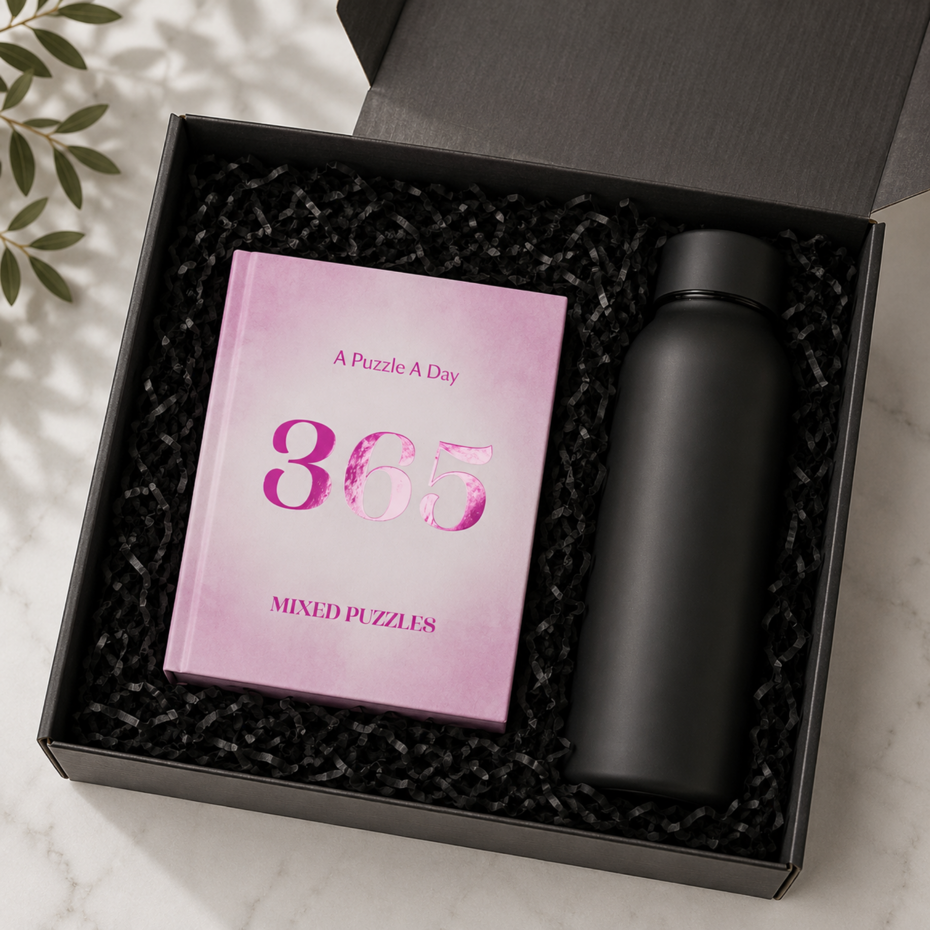 A black gift box filled with black crinkle paper packaging, containing a pink book titled 'A Puzzle A Day 365 Mixed Puzzles' and a matte black bottle, photographed on a light marble surface with leaf shadows.