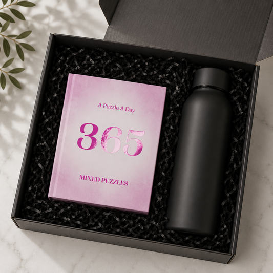 A black gift box filled with black crinkle paper packaging, containing a pink book titled 'A Puzzle A Day 365 Mixed Puzzles' and a matte black bottle, photographed on a light marble surface with leaf shadows.