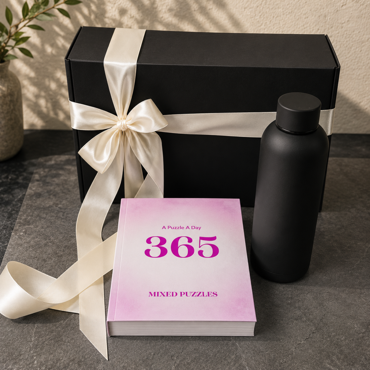 A boxed puzzle gift set on a dark stone surface, featuring a black gift box tied with a cream satin ribbon, a pink book titled "365 Mixed Puzzles," and a matte black bottle.