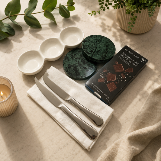 Table setting with ceramic bowls, knives, a box of chocolates, and a candle on a light surface.