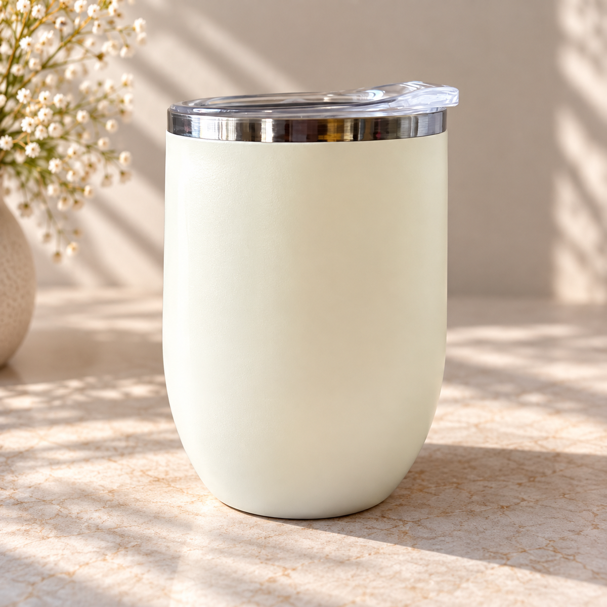 A matte cream-colored insulated travel tumbler with a clear plastic lid and a shiny silver metal rim, set on a light stone surface with soft shadows and baby's breath flowers in the background.
