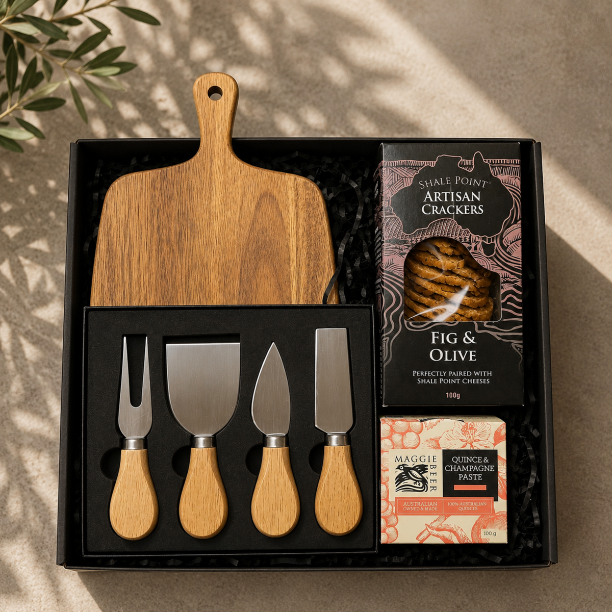 A gift set in a black presentation box featuring a wooden cutting board, a 4-piece wooden-handled cheese knife set, a box of Shire Point Artisan Crackers, and a packet of Margelle Quinoa & Cranberry Paste, arranged on a neutral textured surface with leaf shadows.
