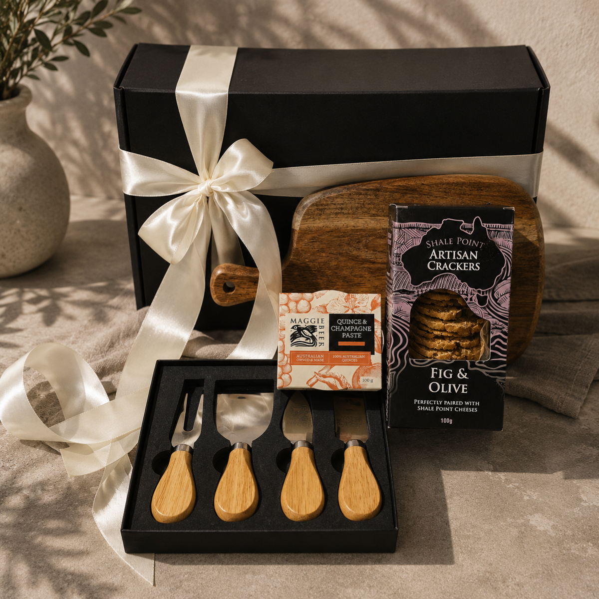 A gourmet gift set featuring a black presentation box with a white satin ribbon, a wooden cutting board, a set of four cheese knives with light wood handles, a box of Shale Pony artisan crackers, and a small bag of Magelli olive oil, arranged on a textured beige fabric.