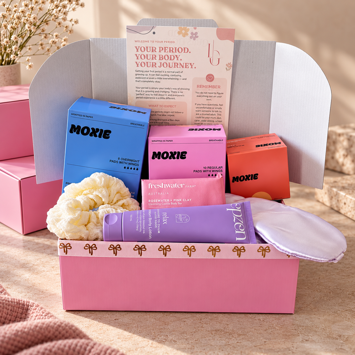 A pink Moxie self-care gift box set arranged in an open pink box on a beige surface. Visible items include a blue Moxie box, a pink Moxie box, an orange Moxie box, a purple open tube, a pale yellow scrunchie, and a white eye mask.