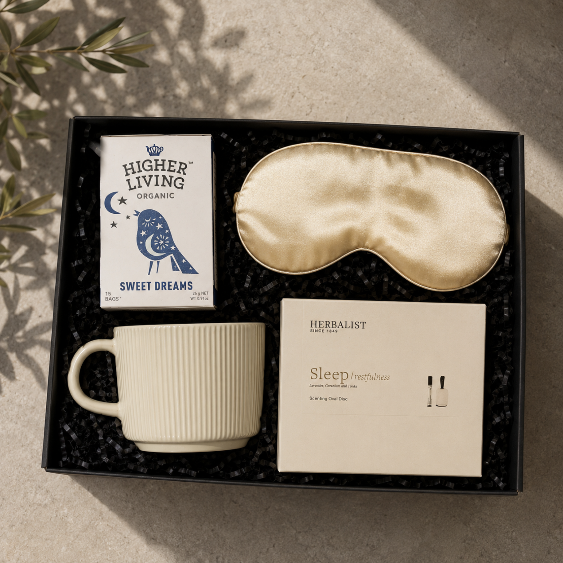A curated gift set in a black presentation box on a sunlit stone surface. The set includes a cream ribbed ceramic mug, a satin sleep mask, a card from Higher Living Organic, and a Herbalist Sleep Freshness card with two small sample vials.