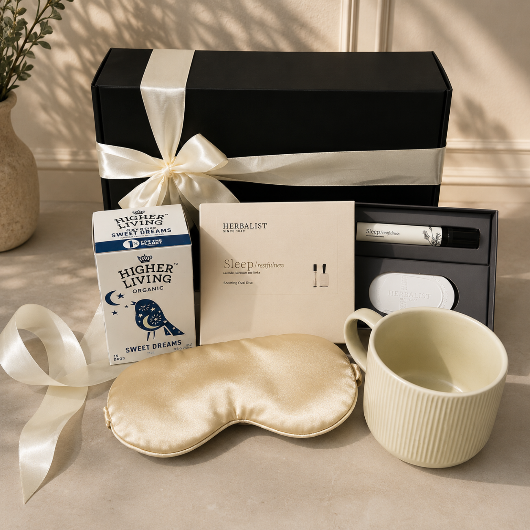 A curated gift set arranged on a beige surface with a black presentation box wrapped in a cream ribbon, a box of Higher Living Organic Sweet Dreams coffee, a Herbalist Sleep Prompts card, a cream sleep mask, a ribbed cream mug, and a small skincare tube in a black case.