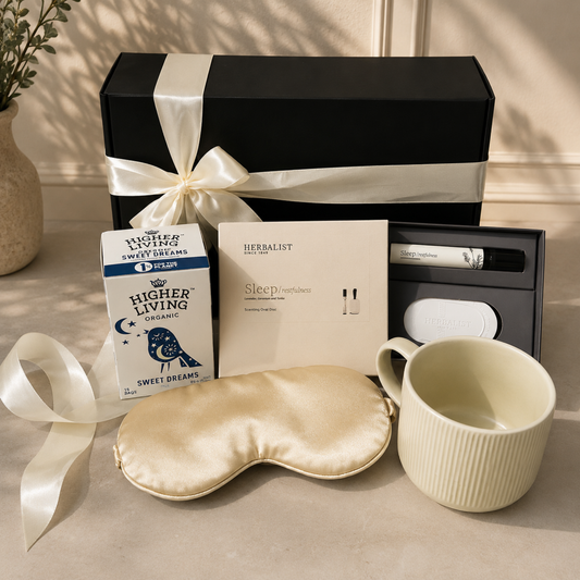 A curated gift set arranged on a beige surface with a black presentation box wrapped in a cream ribbon, a box of Higher Living Organic Sweet Dreams coffee, a Herbalist Sleep Prompts card, a cream sleep mask, a ribbed cream mug, and a small skincare tube in a black case.