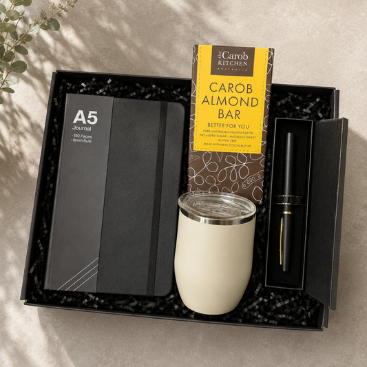 A curated gift set in a black presentation box on a neutral textured surface. The set includes an A5 journal, a Carob Kitchen almond bar box, a black pen in a sleeve, and a cream-colored insulated tumbler with a clear lid.
