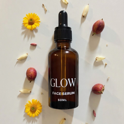 Brown glass bottle labeled 'GLOW FACE SERUM' surrounded by flowers on a light background