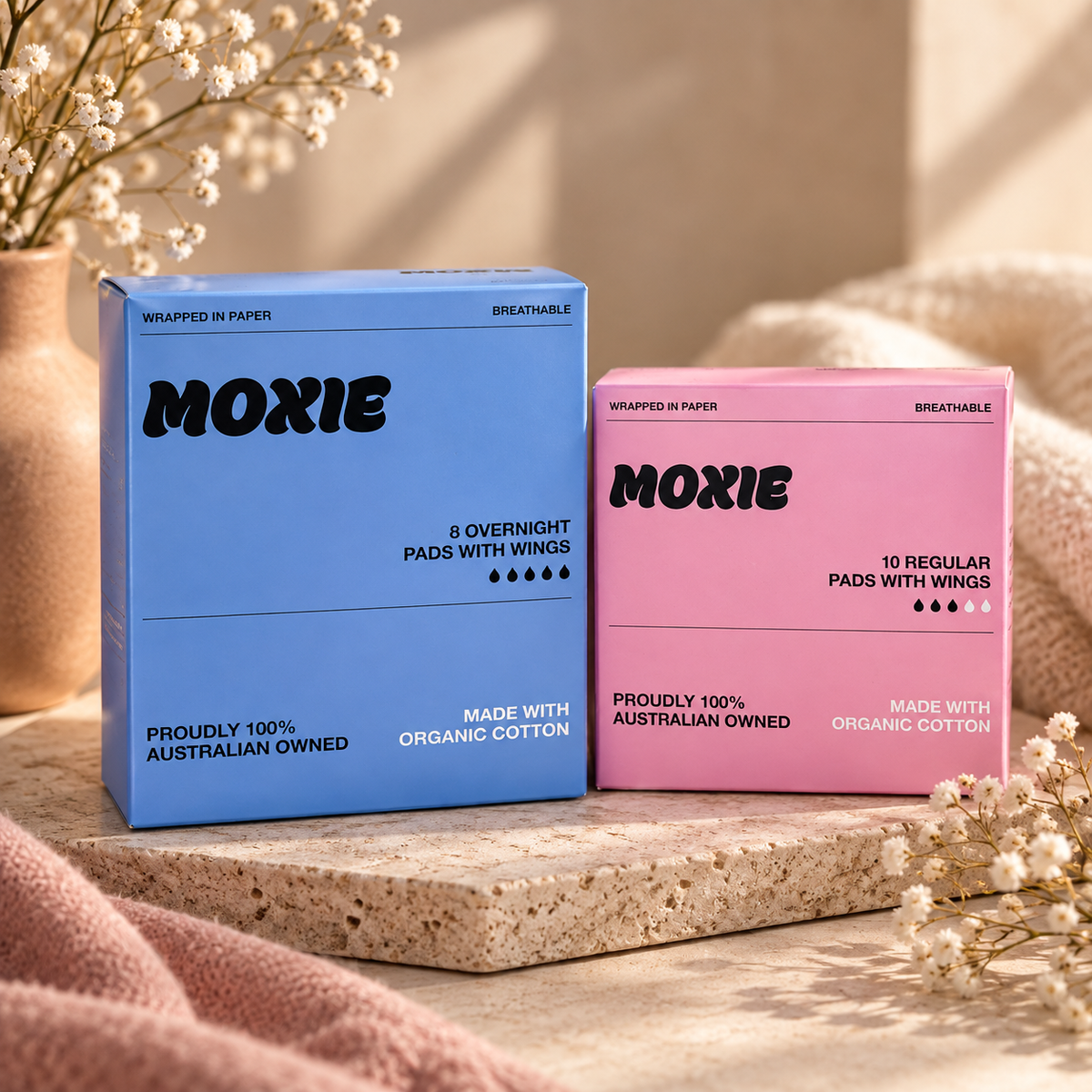 Two Moxie sleep pads boxes, one blue for overnight pads and one pink for regular pads, displayed on a stone slab with soft pink fabric and dried flowers.