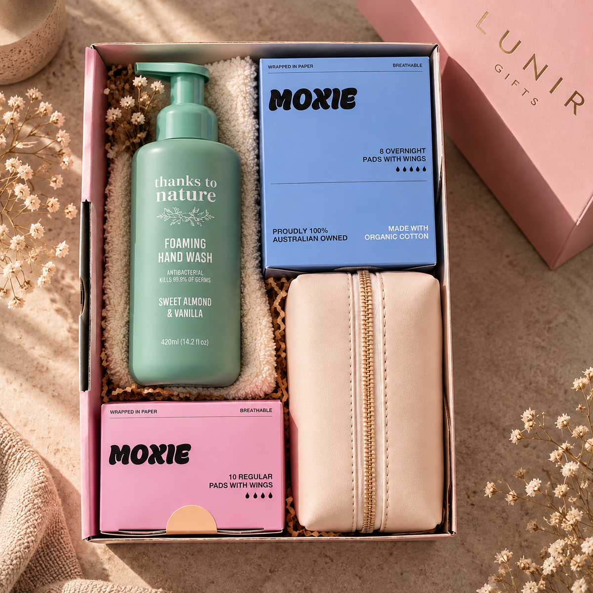 A curated gift box set from Lunir Gifts featuring a green pump bottle of thanks to nature Foaming Hand Wash, two boxes of Moxie pads (one blue, one pink), and a beige zippered pouch, arranged in a pink gift box on a textured beige fabric background.