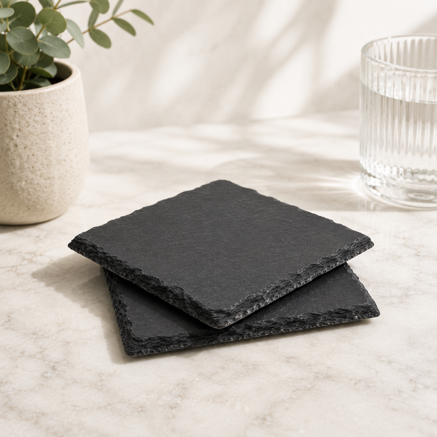 Set of black slate coasters on a light surface with a plant and glass in the background