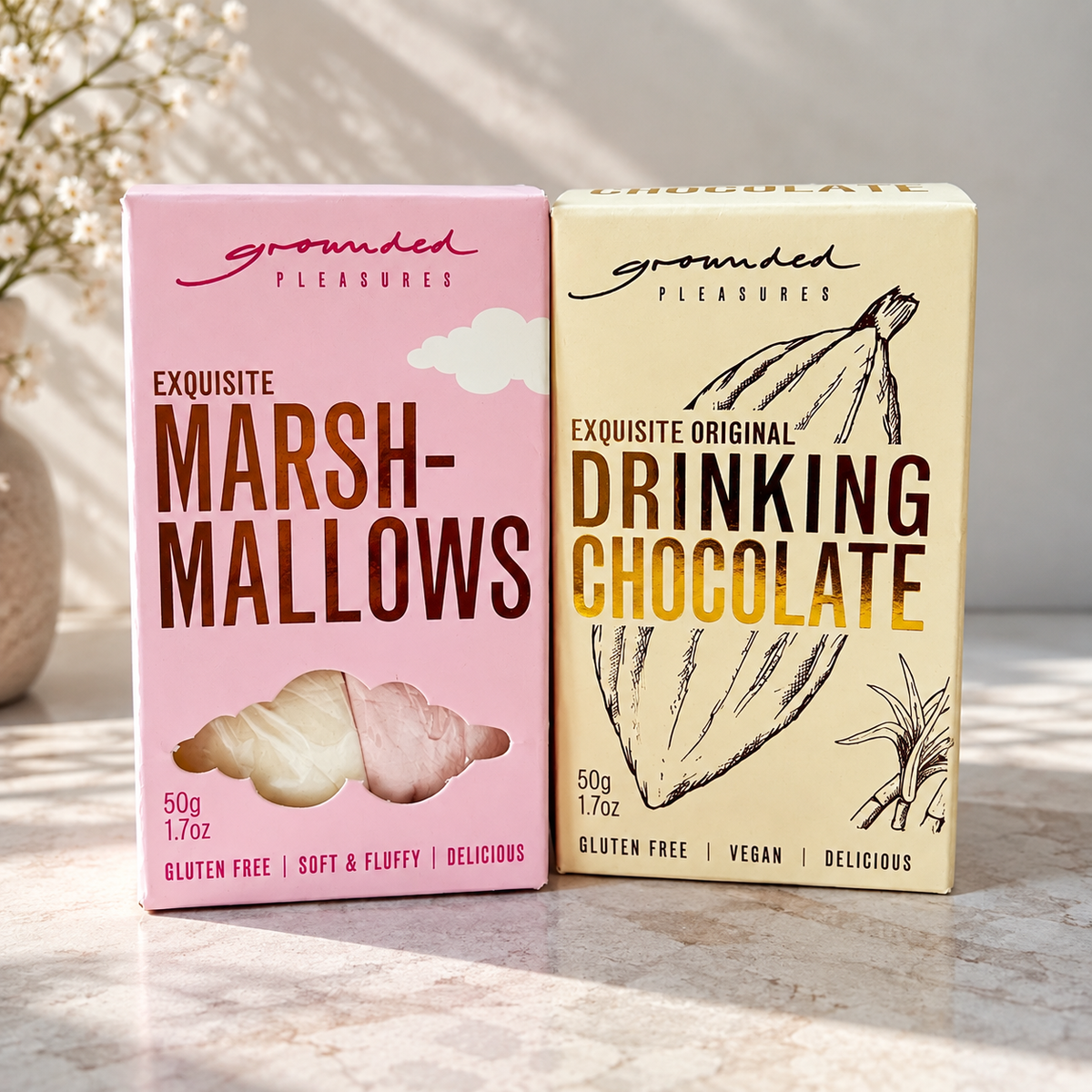 Two boxed chocolate products from Grounded Pleasures on a light marble surface: a pink box of Exquisite Marshmallows and a cream-colored box of Exquisite Original Drinking Chocolate, with soft natural light and blurred dried flowers in the background.