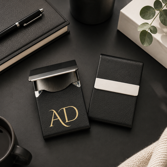 Two black card holders with gold initials on a dark surface with a pen, notebook, and plant.