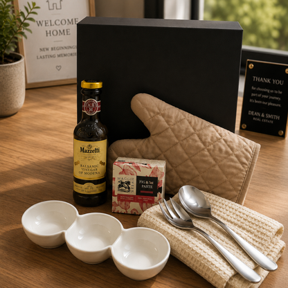 Bottle of Mazzetti vinegar, a box of fig & nut paste, a quilted oven mitt, and cutlery on a wooden surface.