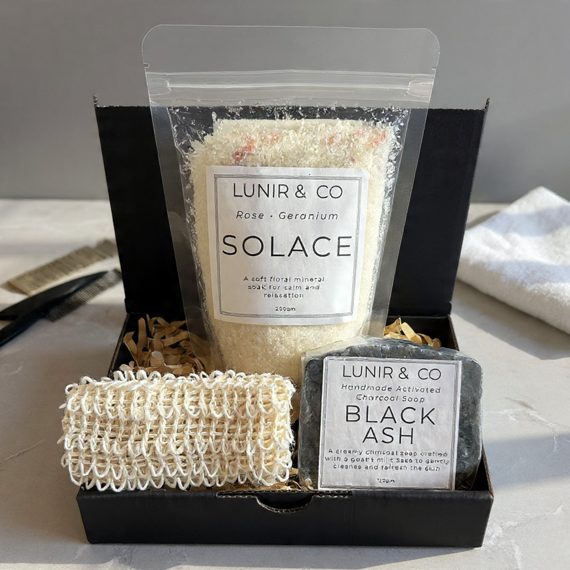 LUNIR & CO bath and body products including a bag labeled 'SOLACE', a soap labeled 'BLACK ASH', and a loofah in a black box.