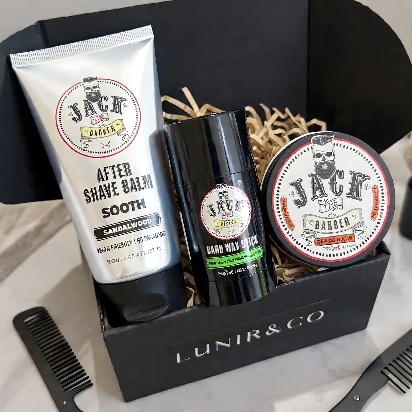 Men's grooming products including an after-shave balm, deodorant stick, and pomade in a branded box on a marble surface.