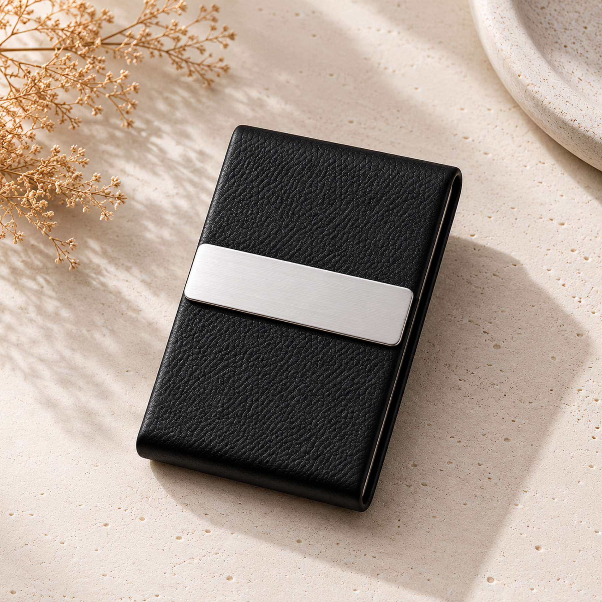 Black leather cardholder with silver accent on a light surface with dried flowers.