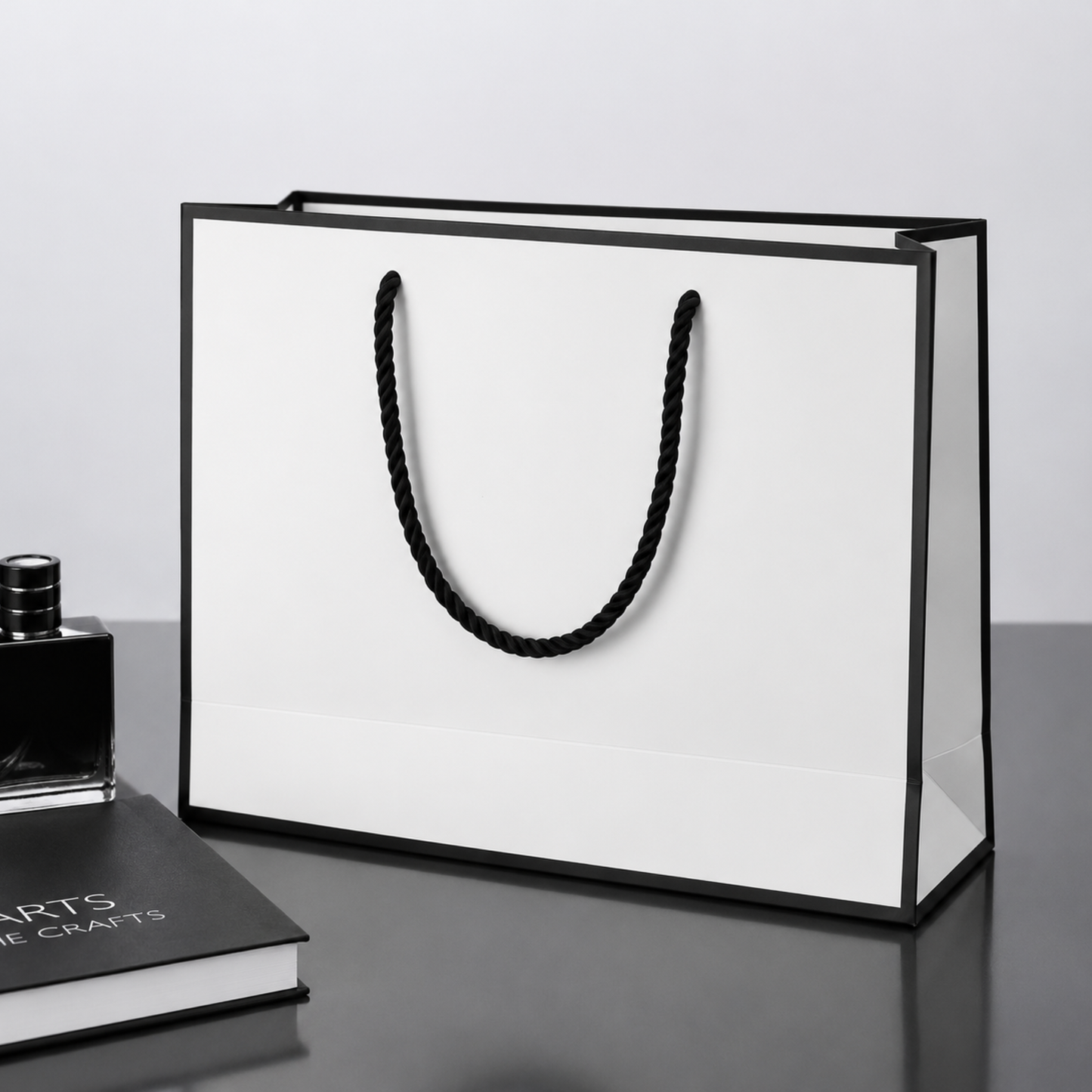 White gift bag with black handle on a gray surface next to a book and bottle.