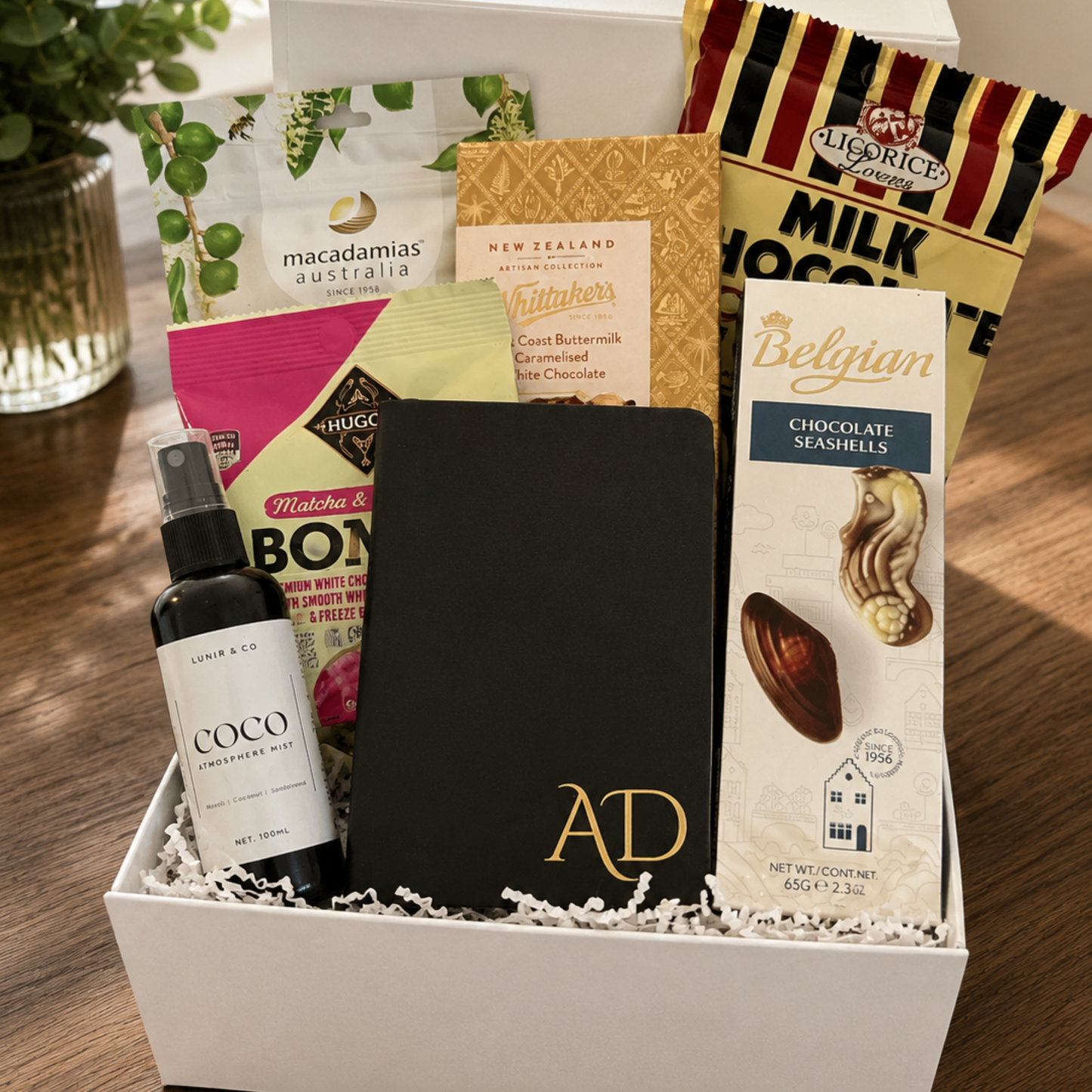 Gift box with various items including a bottle, a book, and a chocolate bar on a wooden surface.