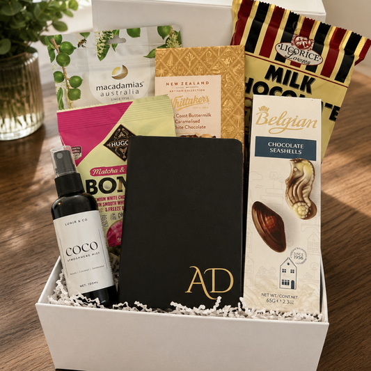 Gift box with various items including a bottle, a book, and a chocolate bar on a wooden surface.