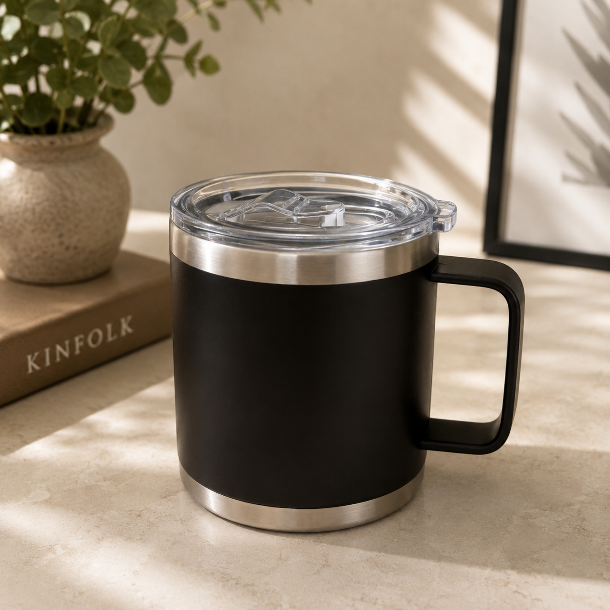 Black insulated mug with silver base and lid on a light surface, next to a plant and book.