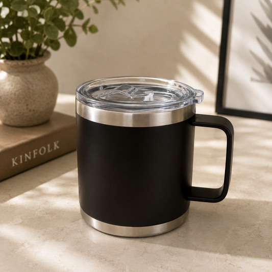 Black insulated mug with silver base and lid on a light surface, next to a plant and book.