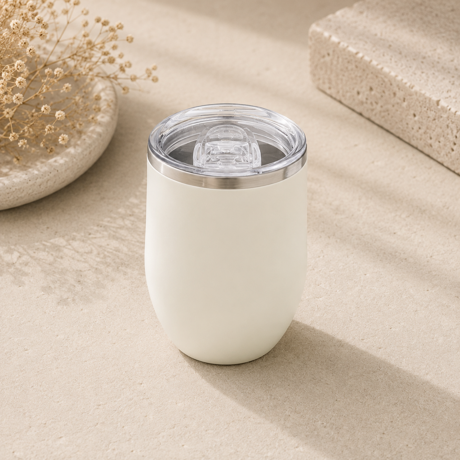 White tumbler with a clear lid on a beige surface with dried flowers and a stone.