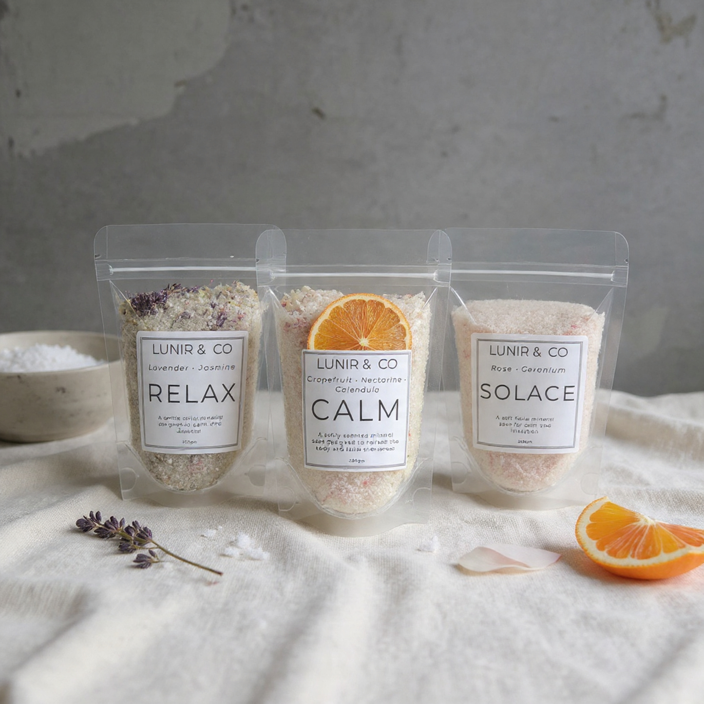Three packages of LUNIR & CO bath salts on a textured surface with oranges and lavender.